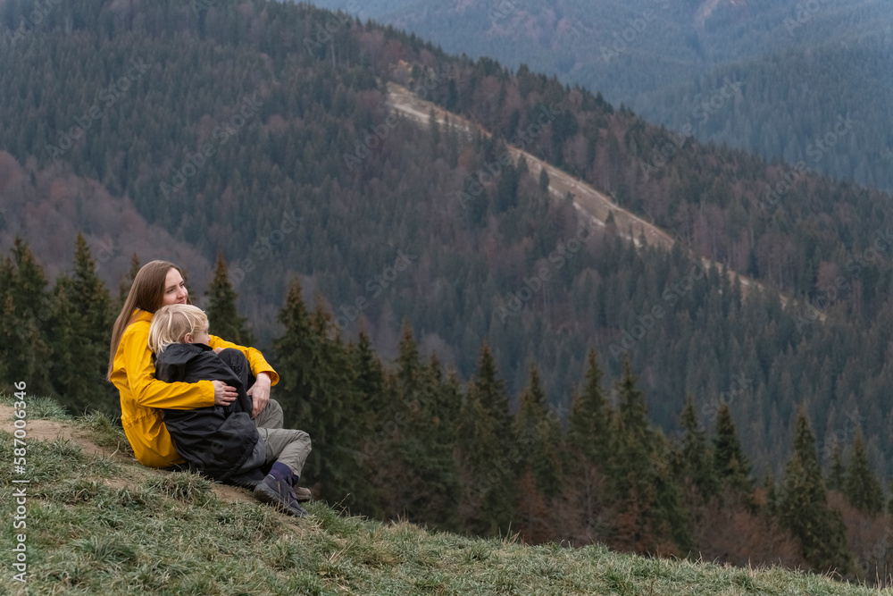Young mother with son sit on hillside. Mother and son rest and sit in an embrace on mountains background.