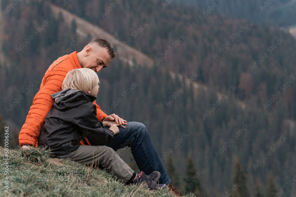 Father and his son sit on slope and look at the mountains. Family trekking with children in forest