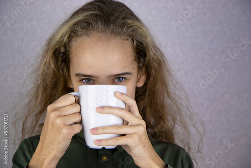 A teenage girl is holding a white cup in her hands, hiding her face behind it. Close-up.