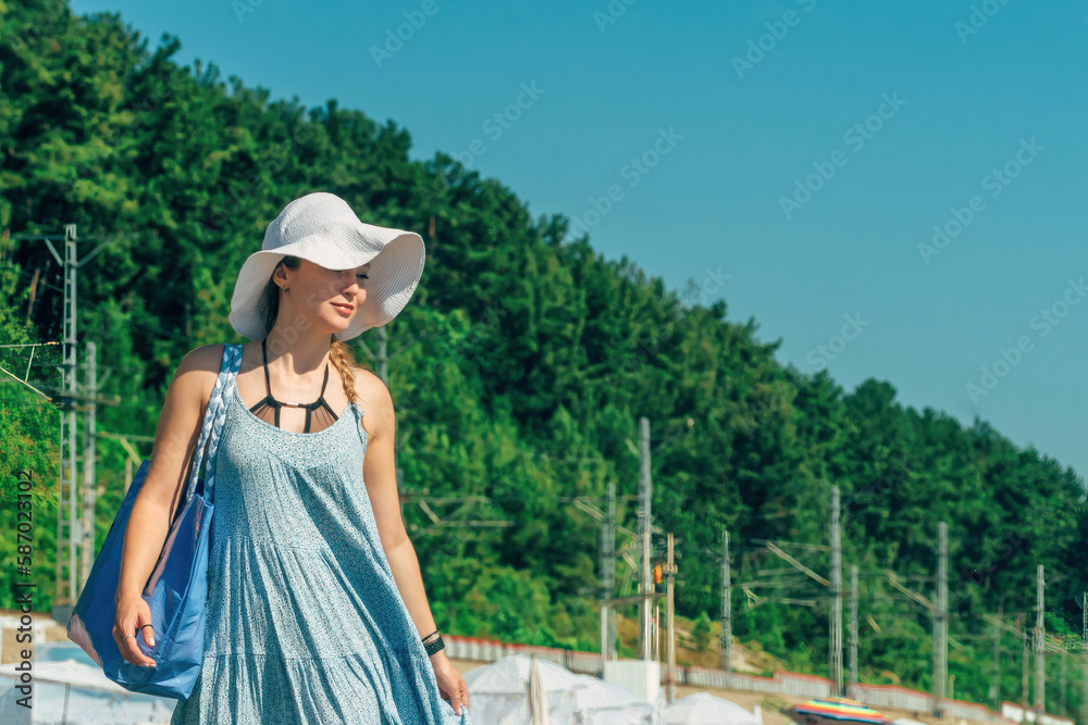 young beautiful woman traveler in a blue summer sundress, sun hat and with a beach bag walks along the seashore during a summer vacation, copy space