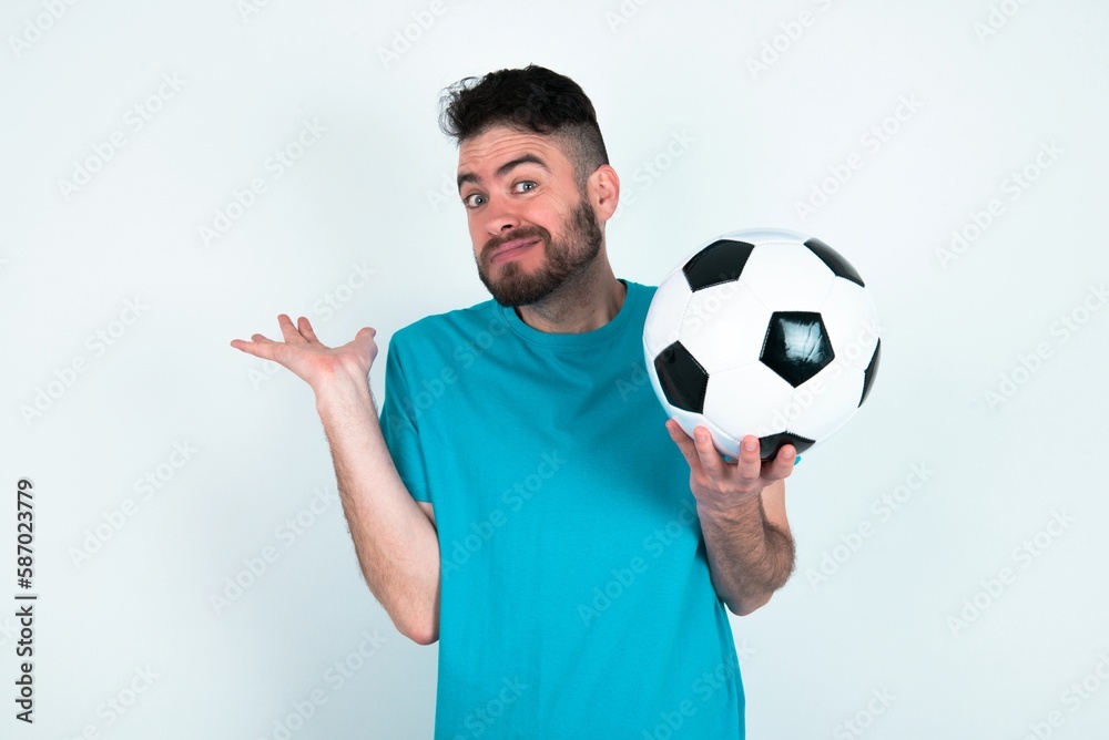 Clueless Young man holding a ball over white background shrugs shoulders with hesitation, faces doubtful situation, spreads palms, Hard decision