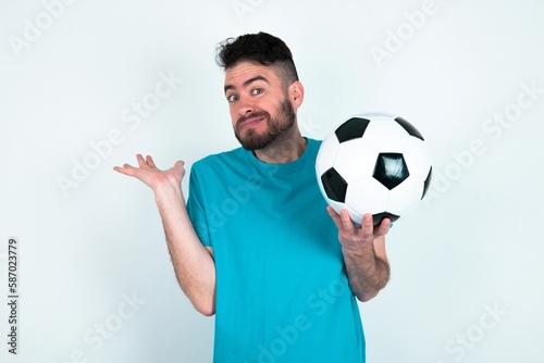 Clueless Young man holding a ball over white background shrugs shoulders with hesitation, faces doubtful situation, spreads palms, Hard decision © Jihan
