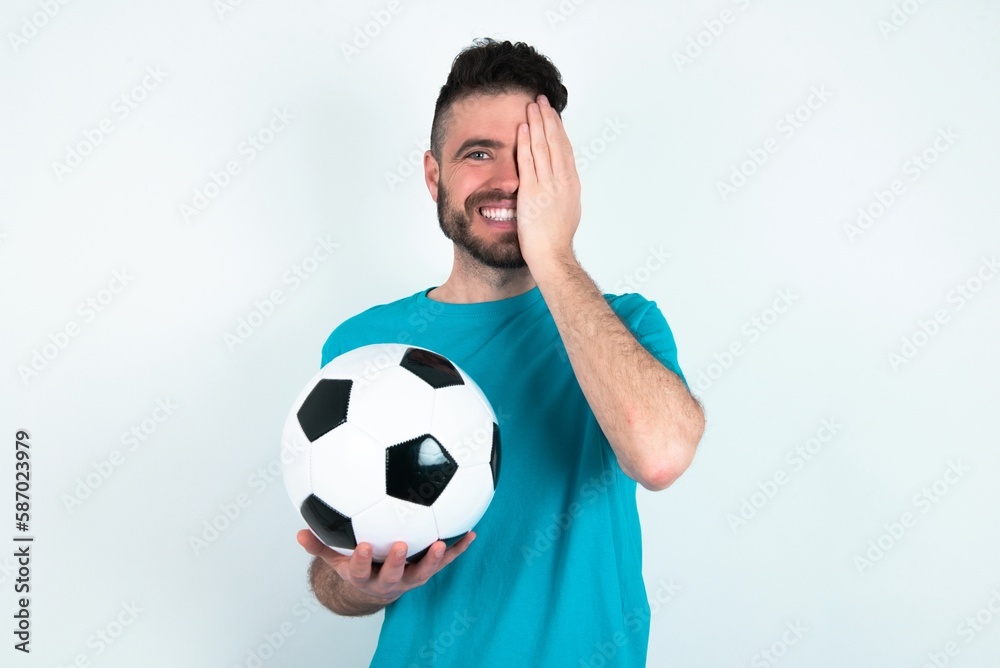 Young man holding a ball over white background covering one eye with her hand, confident smile on face and surprise emotion. Young man holding a ball over white background covering one eye with her hand, confident smile on face and surprise emotion.
