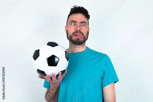Young man holding a ball over white background showing grimace face crossing eyes and showing tongue. Being funny and crazy