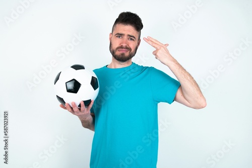 Unhappy Young man holding a ball over white background makes suicide gesture and imitates gun with hand, curves lips, keeps two fingers on temple