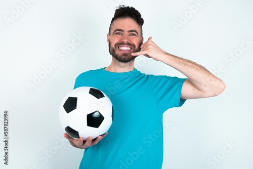 Young man holding a ball over white background smiling cheerfully and pointing to camera while making a call you later gesture, talking on phone