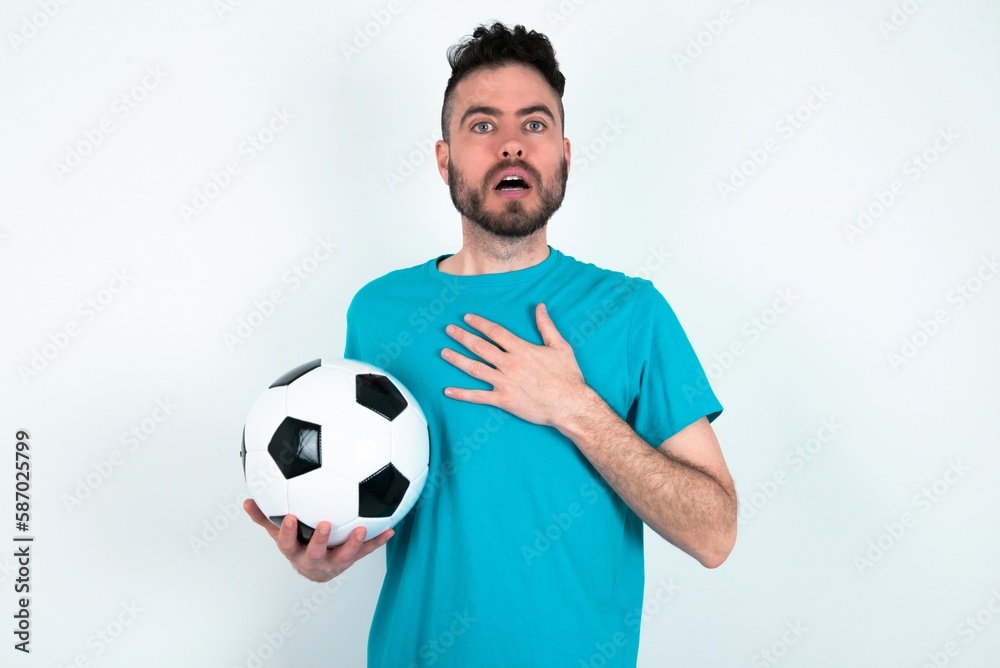 Scared Young man holding a ball over white background looks with frightened expression, keeps hands on chest, being puzzled to notice something strange, People, hush reaction and emotions. Scared Young man holding a ball over white background looks with frightened expression, keeps hands on chest, being puzzled to notice something strange, People, hush reaction and emotions.