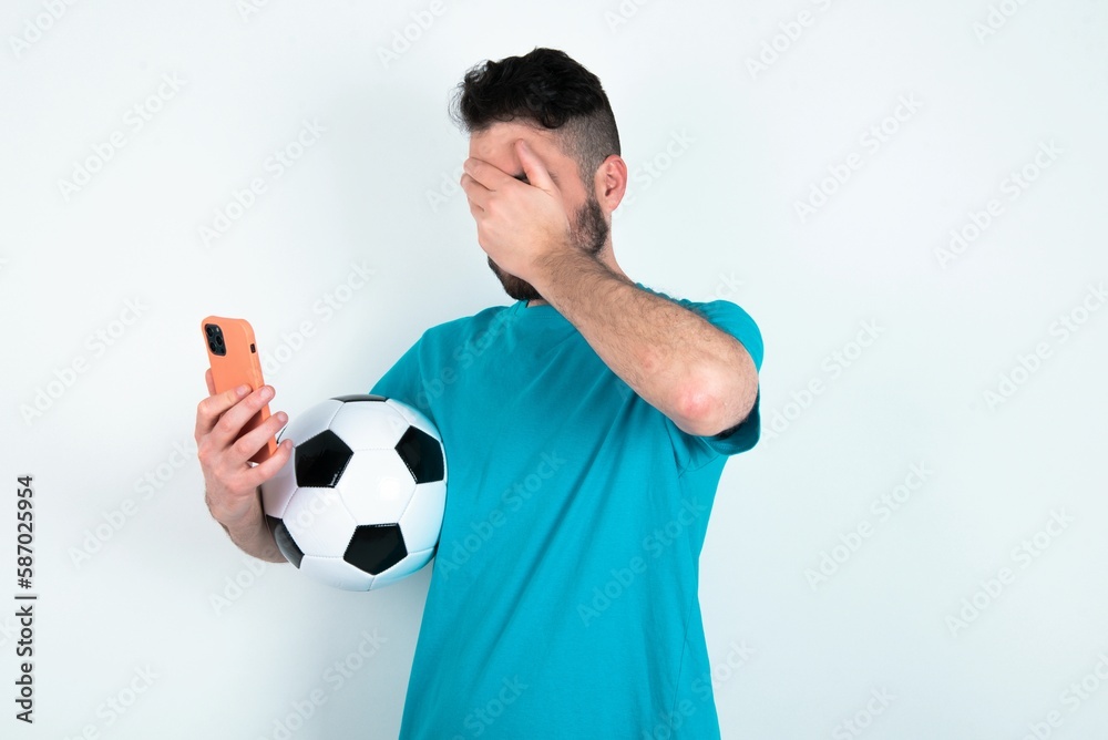 Young man holding a ball over white background looking at smart phone feeling sad holding hand on face.