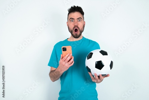 Shocked Young man holding a ball over white background opens mouth hold phone reading advert unbelievable big shopping prices