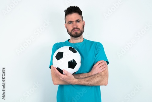 Gloomy dissatisfied Young man holding a ball over white background looks with miserable expression at camera from under forehead, makes unhappy grimace