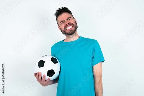 Positive Young man holding a ball over white background with overjoyed expression closes eyes and laughs shows white perfect teeth