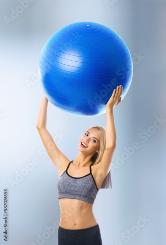 Portrait of happy excited blond woman in sportswear with fitball, standing on blurred modern gym center background. Fitness, exercising and fit gym concept.