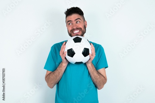 Happy Young man holding a ball over white background anticipates something awesome happen, looks happily aside, keeps hands together near face, has glad expression.