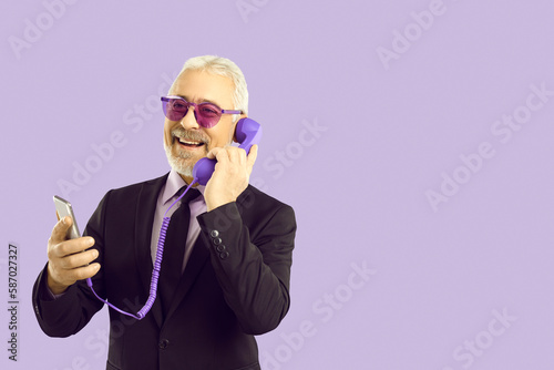 Funny mature bearded man in suit isolated on purple studio background talk on cellphone using corded headset. Smiling senior male have smartphone call on landline set. Communication.