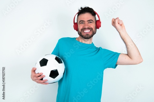 Carefree Young man holding a ball over white background with toothy smile raises arms dances carefree moves with rhythm of music listens music from playlist via headphones