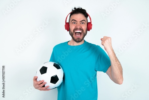 Emotional Young man holding a ball over white wall exclaims loudly feels like winner raises clenched fists keeps mouth opened wears stereo headphones on ears makes yes gesture, listens favorite music