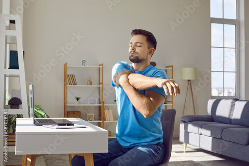 Man stretches during work break. Young man in blue T shirt sitting by desk with laptop computer in home office and doing relaxing exercise to prevent muscle pain, discomfort and stiffness in shoulders