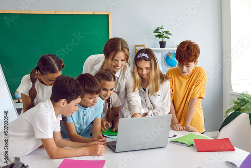 Billede på lærred Smart children pupil boys and girls stand in front of laptop with woman teacher and view media material or online presentation during lesson in primary school classroom
