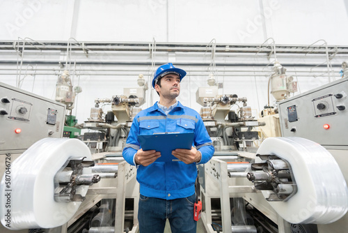 A white European male engineer with a smart look in a plastics factory. Around there are machines working. holding a listnote and a walkie-talkie Wear uniforms and helmets.