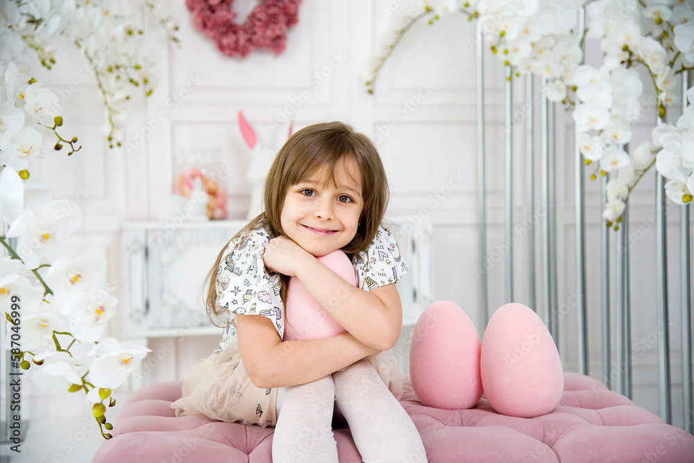 little funny cute smiling brunette girl sitting on oink sofa and playing with three big pink toy easter egg in room with white orchids little funny cute smiling brunette girl sitting on oink sofa and playing with three big pink toy easter egg in room with white orchids
