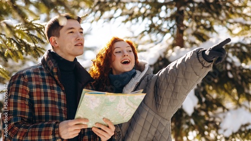 A red-haired woman standing next to her boyfriend points her hand somewhere towards the exit from the forest. Young people stand in the winter forest with a map in their hands and look somewhere.