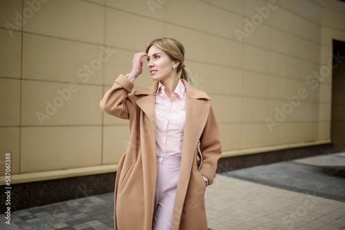 Portrait of a smiling young caucasian woman in a coat fixing her hair with her hand looking away, outside. Stylish woman posing on the street. The concept of management, recreation, photoshoot