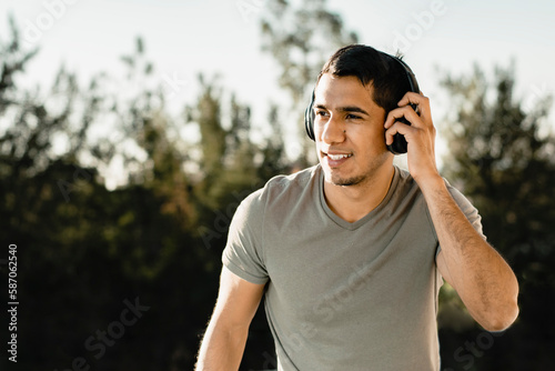 Persona masculina escuchando música con cara sonriente y manos sosteniendo los audífonos con arboles detrás de él