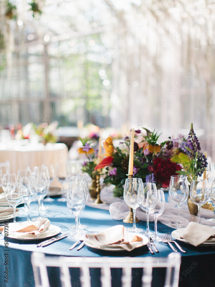 Wedding banquet table setting in blue and white. In the foreground are white plates with napkins, wine glasses, knives and forks. Behind bright flowers. Drapery with tulle. Wedding banquet table setting in blue and white. In the foreground are white plates with napkins, wine glasses, knives and forks. Behind bright flowers. Drapery with tulle.