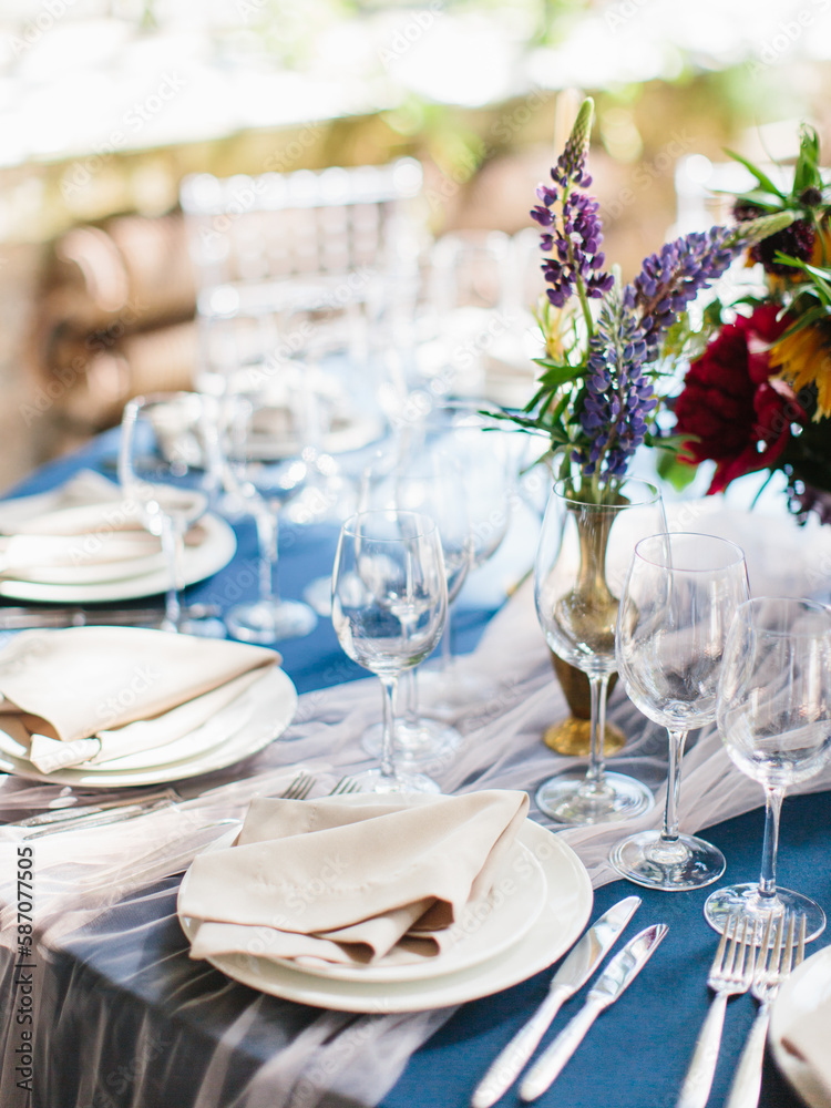 Wedding banquet table setting in blue and white. In the foreground are white plates with napkins, wine glasses, knives and forks. Behind bright flowers. Drapery with tulle.