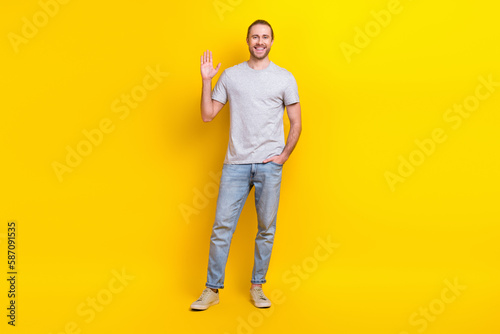 Full size photo of good mood friendly man wear gray t-shirt jeans hand in pocket waving palm say hi isolated on yellow color background