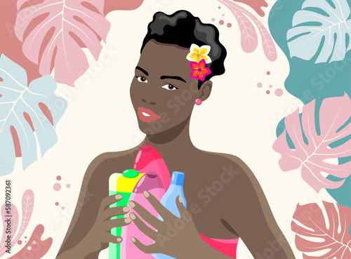 Beautiful dark-skinned smiling girl with spa products in her hands and flowers in short black hair on a light background with tropical leaves