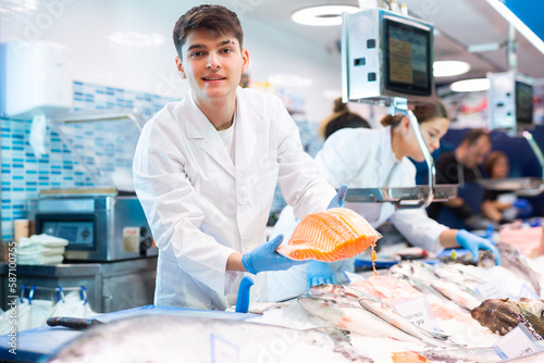 Proficient shop assistant in work clothes holding in hands salmon in fish shop