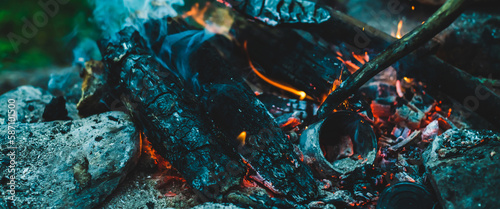 Vivid smoldered firewoods burned in fire close-up. Atmospheric warm background with orange flame of campfire and blue smoke. Unimaginable full frame image of bonfire. Burning logs in beautiful fire.