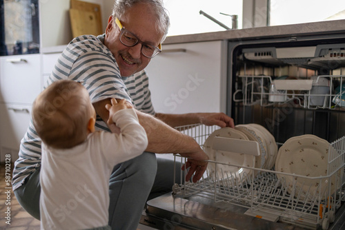 Smiling grey haired matured father making dishes near dishwasher with his baby son