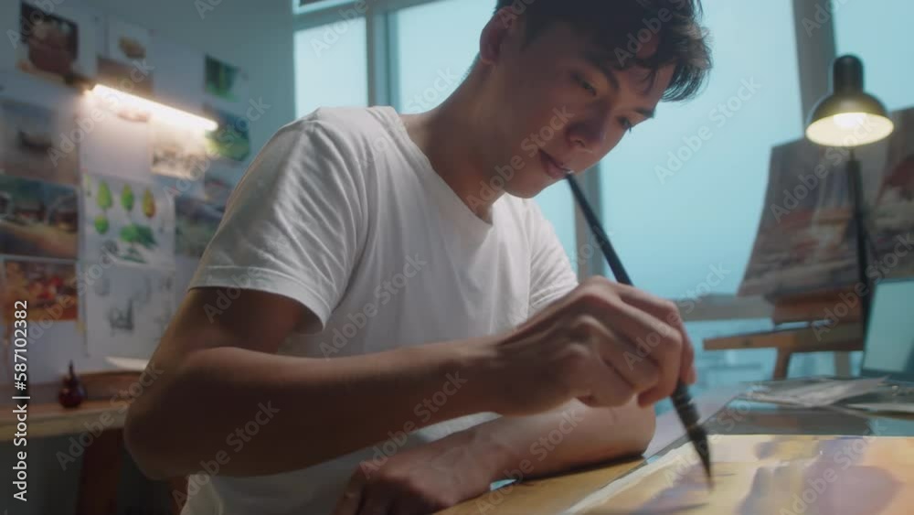 Medium selective focus shot of young Asian man painting picture at desk while working in art studio Medium selective focus shot of young Asian man painting picture at desk while working in art studio