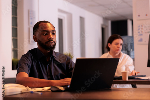 Employee using corporate laptop in coworking space office, worker sitting at workplace desk. African american businessman working on startup project report on computer at night