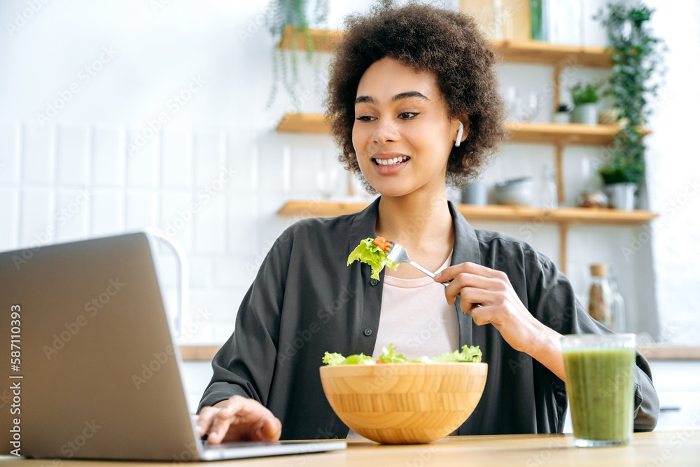 Healthy lifestyle concept. Modern positive african american young woman, sitting at table in kitchen, eating fresh vegetable salad, watching healthy food or home workout videos on laptop, smiling