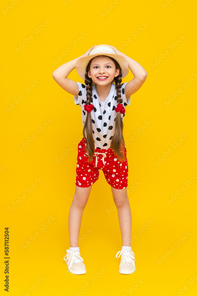 A full-length portrait of a little girl in red shorts, a white T-shirt and a straw hat, holding her hat with both hands and smiling broadly, rejoicing in the summer. Yellow isolated background. A full-length portrait of a little girl in red shorts, a white T-shirt and a straw hat, holding her hat with both hands and smiling broadly, rejoicing in the summer. Yellow isolated background.