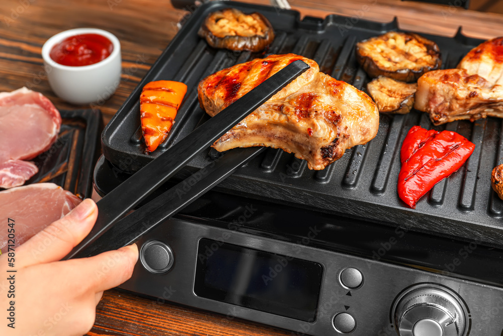 Woman cooking tasty steaks and vegetables on modern electric grill at table, closeup Woman cooking tasty steaks and vegetables on modern electric grill at table, closeup