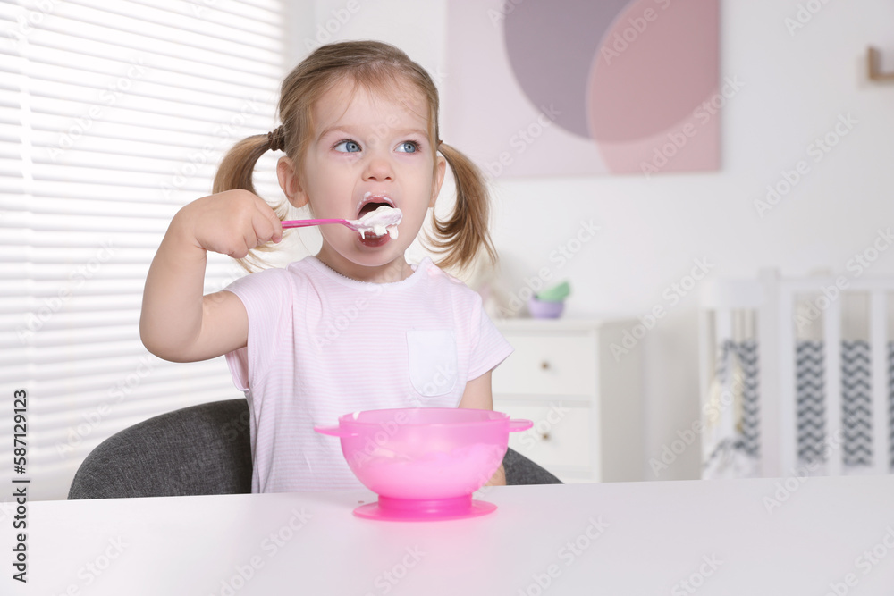 Cute little child eating tasty yogurt from plastic bowl with spoon at white table indoors Cute little child eating tasty yogurt from plastic bowl with spoon at white table indoors