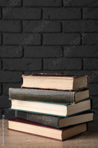 Stack of old hardcover books on wooden table near black brick wall, space for text