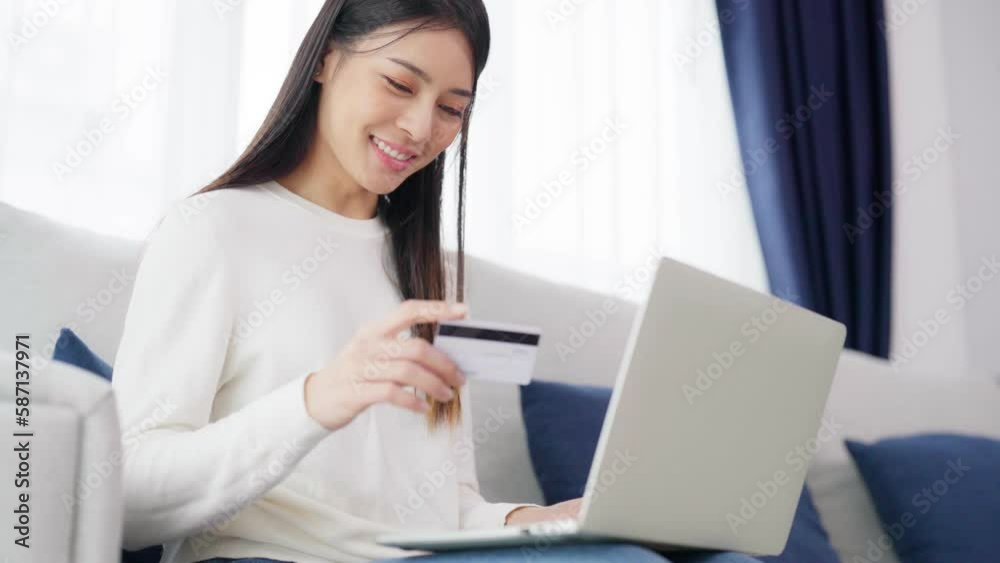 Young asian woman sitting on sofa in living room, makes online banking payments through the internet from bank card on computer laptop. Shopping online on notebook with credit card Young asian woman sitting on sofa in living room, makes online banking payments through the internet from bank card on computer laptop. Shopping online on notebook with credit card