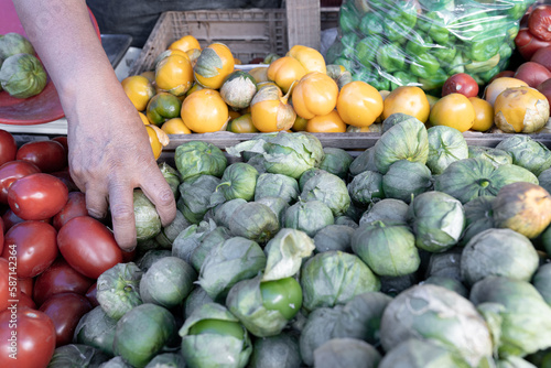 An adult woman is taking a green tomato from a street market stall with yellow, red and green