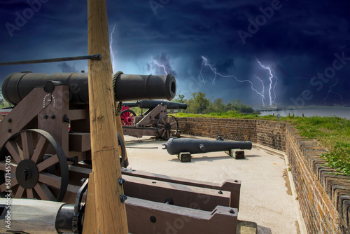 Photography black cannons with a brown wooden base in front of a red brick wall with lush gr