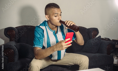 young hispanic soccer fan man sitting drinking a beer while watching a phone