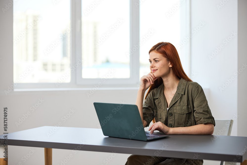 red-haired woman smiling thoughtfully sitting at work at a laptop
