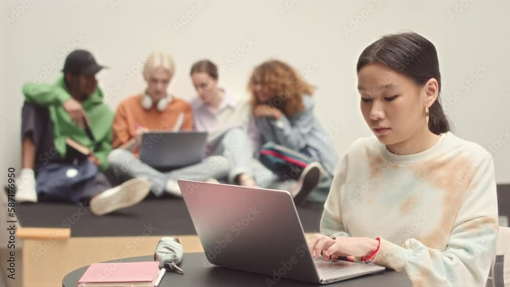 Medium slowmo of young pretty Asian female student studying on laptop at college while her classmates sitting on floor and chatting in blurred background Medium slowmo of young pretty Asian female student studying on laptop at college while her classmates sitting on floor and chatting in blurred background