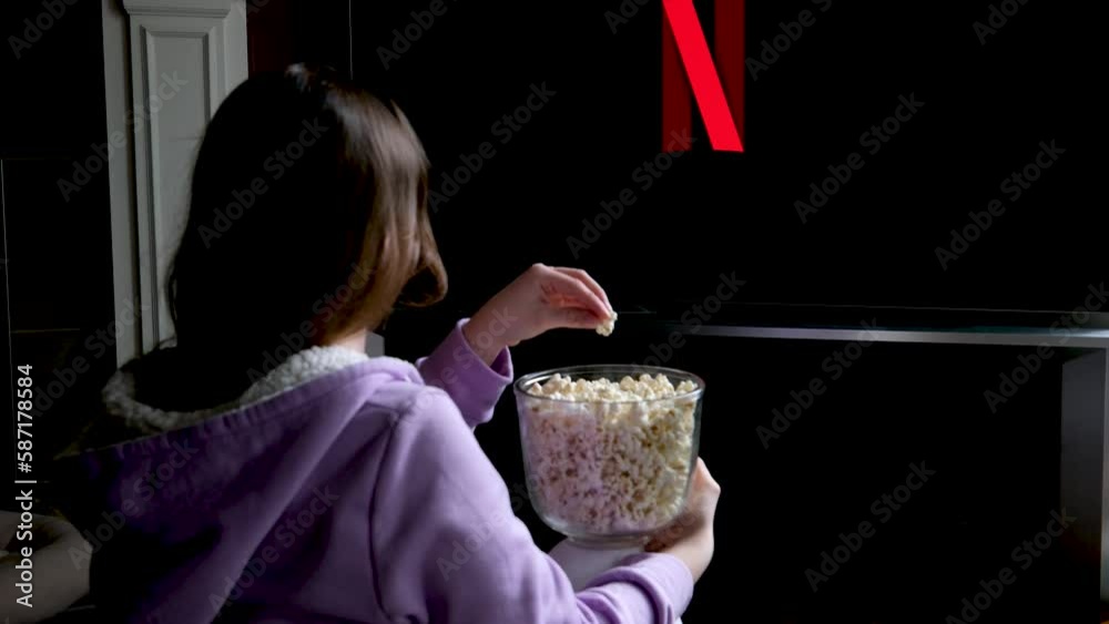 teenage girl sits on floor and watches netflix on big screen of TV screensaver slow motion flashes on black background in red blue in hands of young woman popcorn girl takes it and eats it spellbound