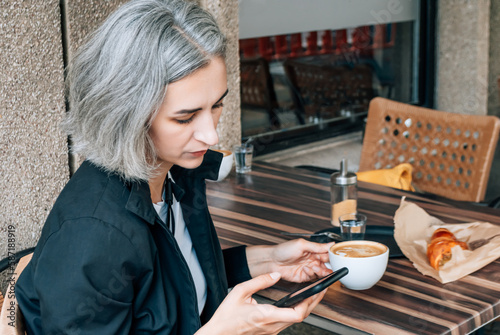 Photography Gray-haired woman sits at street coffee shop and uses a smartphone