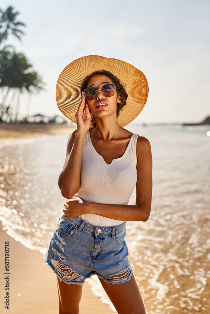 Young african female model in straw hat and sunglasses posing at resort by sea at sunrise. Black woman against scenic rocky green island and ocean tide at dawn. Multiracial model poses on vacation